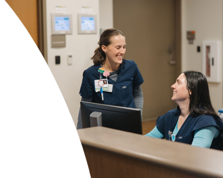 Two nurses in scrubs smiling and collaborating at a nurse's station.